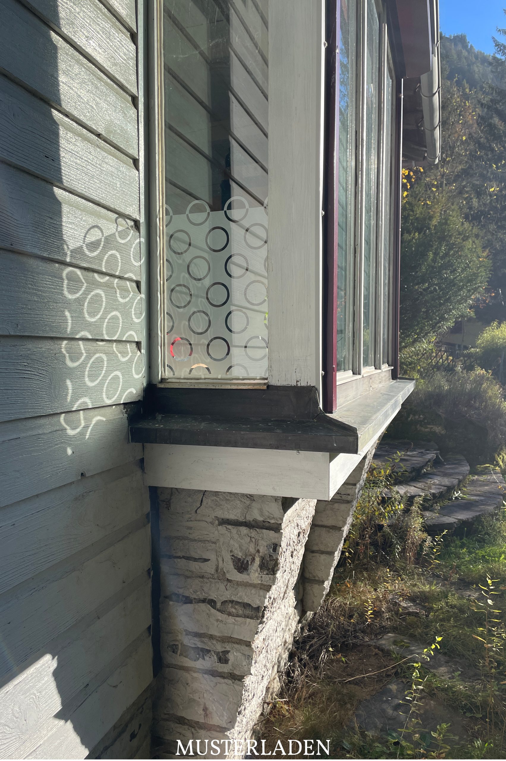 Blick von außen auf Fenster mit Sichtschutzfolie mit geometrischem Muster. Die Sonne erzeugt Schattenspiele des Motivs auf der Holzfassade.